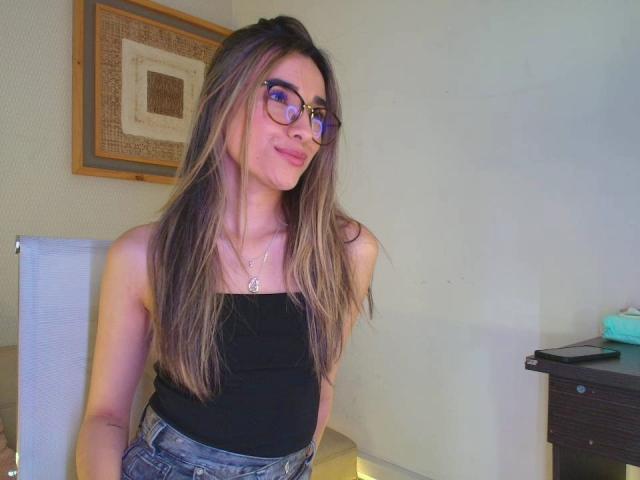 AdharaCooperr mit langen braunen Haaren, Brille, schwarzem Top und blauen Jeans, lächelt leicht, wobei ihre kleinen Brüste sichtbar sind. Sie steht in einem Raum mit einem Holzrahmen und einer weißen Wand..