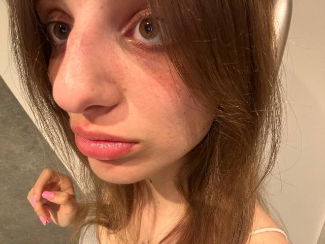 KarieMestler's face, showing a slightly flushed nose, pink lips, brown eyes, and brown hair, with pink-nailbed hand touching her hair. She's wearing a white top.