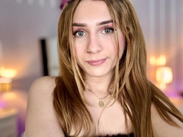 AndyDupont has long brown hair, pink eyeshadow, a nose ring, and a necklace with a circular pendant, smiling at the camera with her tits partially visible.