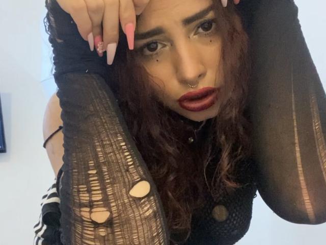 SweetSolei69, com cabelo escuro e ondulado, piercing no nariz e batom vermelho, vestindo uma blusa de malha preta com furos, com as mãos no cabelo, unhas cor-de-rosa, olhando para cima com os lábios entreabertos..