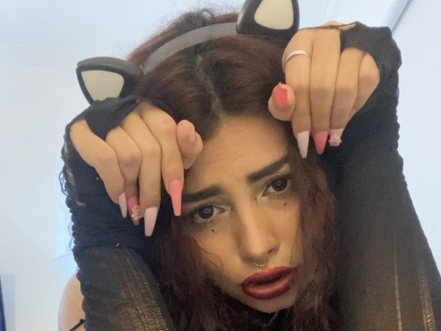 SweetSolei69 with cat ears, dark curly hair, red lipstick, nose ring, and long pink nails, hands on head, looking up with slightly open mouth, wearing black sleeves.