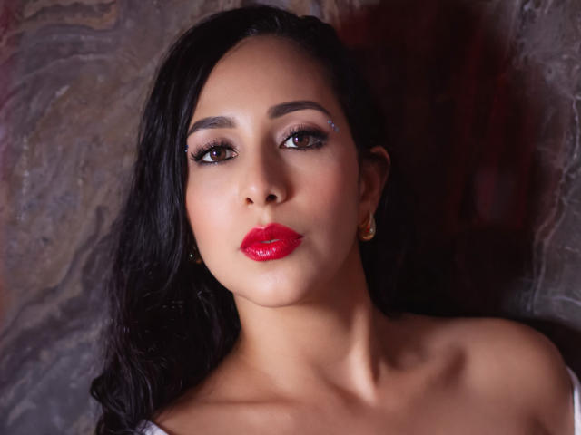 KattyRivera a les cheveux foncés, les yeux marron, du rouge à lèvres, un petit piercing au nez, et des boucles d'oreilles dorées, avec une peau lisse et bronzée, et un visage sérieux et attrayant..