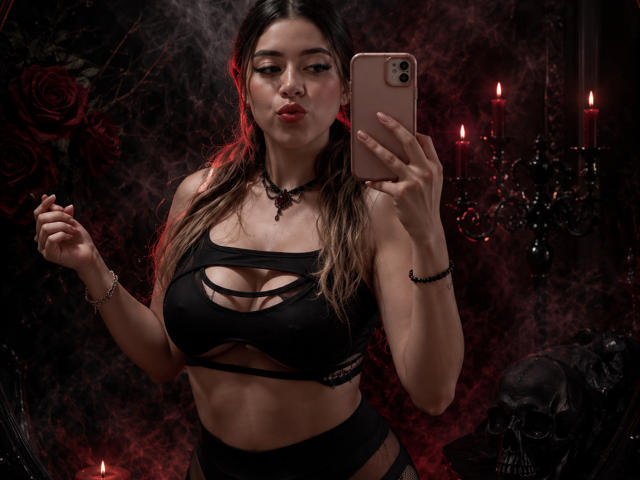 IsabellaFetishism, avec de longs cheveux bruns, des lèvres pulpeuses et un haut noir à bretelles laissant entrevoir ses seins, tenant un téléphone, dans une pièce sombre et gothique avec des bougies rouges et un crâne..