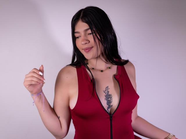 SelenaRios with black hair, wearing a red zip-up top, showing a chest tattoo, holding a bracelet, smiling slightly, necklace with small green pendants.