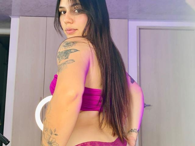 SophiaMarx with long brown hair, purple lace bra, and matching panties, showing tattoos on her arms and back, looking over her shoulder with a slight smile.