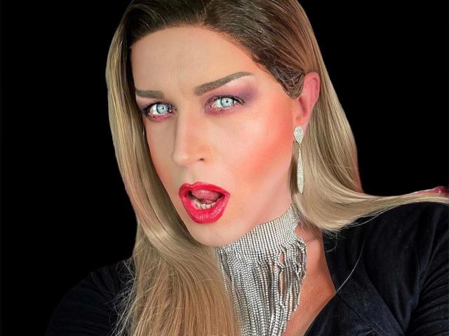 CassyTV, avec ses cheveux blonds, son rouge à lèvres rouge et son expression de surprise. Elle porte un collier scintillant et des boucles d'oreilles en diamant, et a l'air déterminée avec son fard à paupières violet et ses yeux bleu vif..