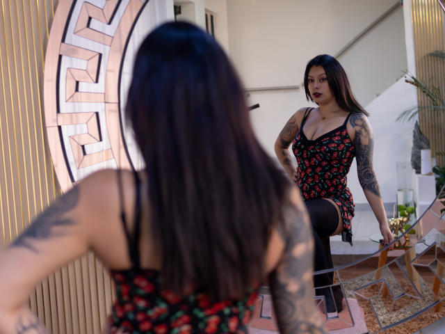 GabyJoy, con i suoi lunghi capelli neri, le braccia tatuate, indossa un abito rosso con fiori e calze nere, e fissa il suo riflesso in uno specchio. Ha il rossetto scuro e un'espressione sicura..