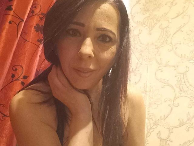 LauraSin with long dark brown hair, brown eyes, and slight smile, holding her neck, topless, showing small tits, in a warmly lit room, wearing small earrings.