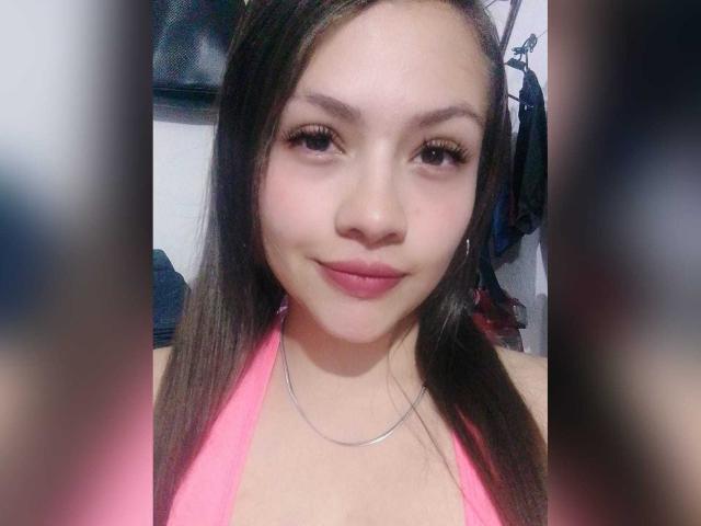 NoaLaurentier with long brown hair, pink halter top, and subtle smile, showing small tits, pink lips, and brown eyes, wearing a silver necklace.