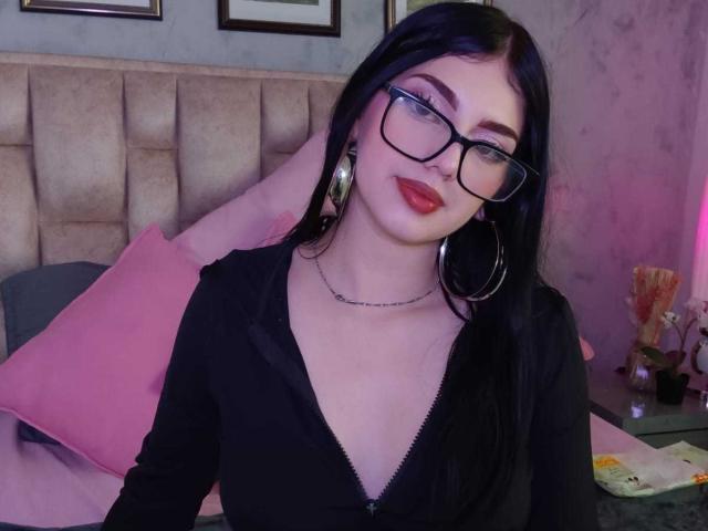 Isabella Rossi, avec des cheveux noirs, des lunettes, du rouge à lèvres rouge, de grandes boucles d'oreilles, un haut noir à fermeture éclair, une peau pâle, assise sur un lit, avec un oreiller rose, une tête de lit grise, un petit collier, souriante..