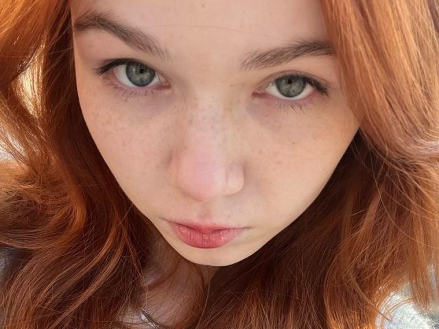 KatiRokisky's face, with pale skin, freckles, and red wavy hair, staring intensely at the camera. Her green eyes are centered, and her pink lips are slightly pursed.