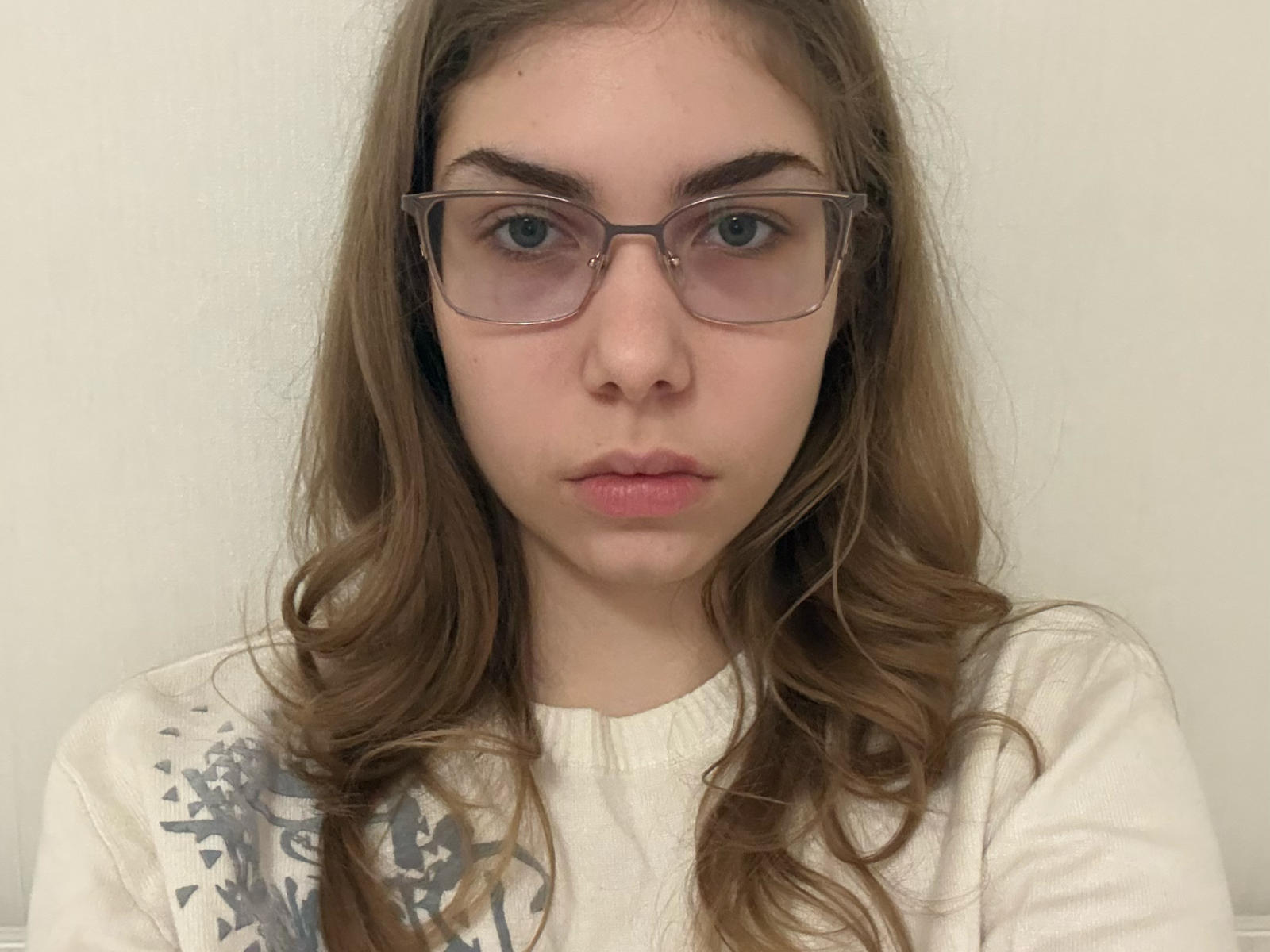 EthelynPerrot with long brown wavy hair, wearing glasses, white sweater, and pinkish lips, staring directly at camera, neutral expression, soft light. Boobs not visible, focus on face.