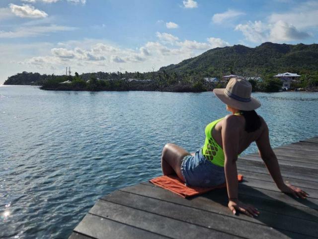 EmaFoxy mit gebräunter Haut, einem grünen, neonfarbenen Oberteil, Jeansshorts und einem Sonnenhut, sitzt auf einem Steg, mit dem Rücken zur Kamera, und blickt auf das ruhige, blaue Wasser. Sie trägt ein orangefarbenes Handtuch..