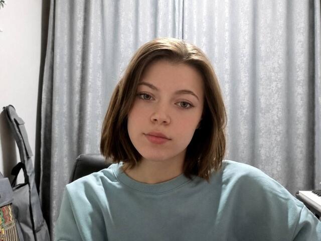 JennellMatacale with short brown hair, wearing a teal sweater, has a neutral expression, sitting in front of gray curtains, with a black bag to the left.