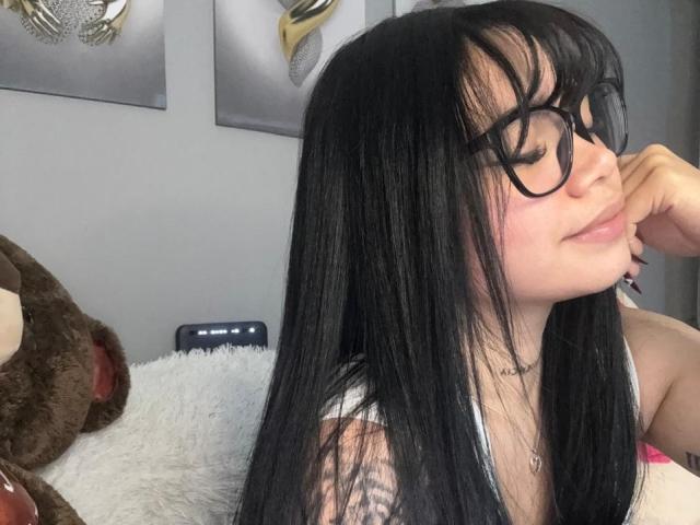 LenaLegrand with black hair, glasses, and a nose piercing, smiling slightly, touching her chin, with a white stuffed animal and brown teddy bear in the background.