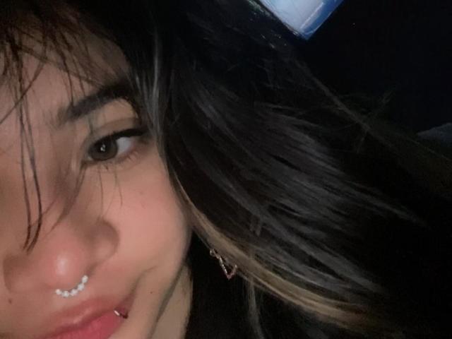 AmaraKazumi's face, showing her nose ring, pierced lip, and long black hair with blonde highlights, slightly messy, dark eyes looking at the camera.