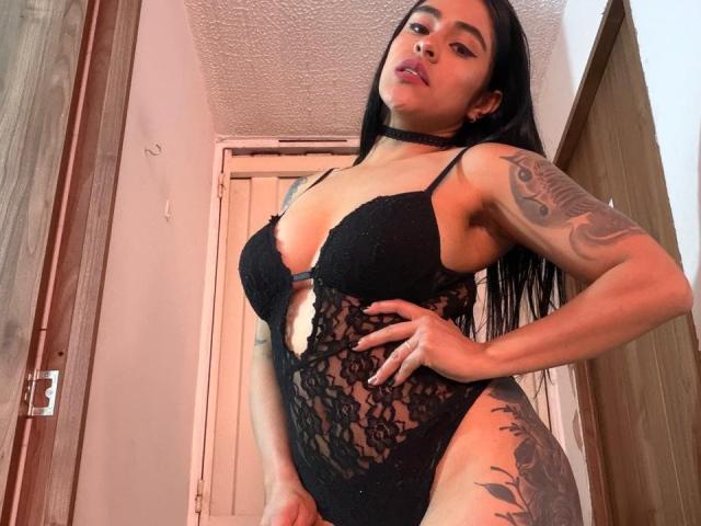 StellaBeckker with long black hair, wearing a black lace bodysuit, choker, and tattoos, standing in a room, hands on hips, showing her tits and a small cutout in the lace.