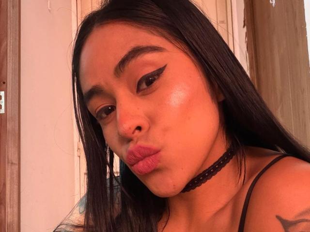 StellaBeckker with long black hair, bold eyeliner, shiny skin, pouty lips, black choker, and a tattoo on her right shoulder, looking at the camera with a slightly puffy face.