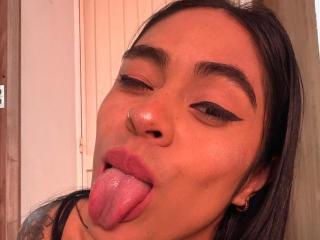 StellaBeckker with brown skin, black eyeliner, and nose ring, sticking out her pink tongue, eyes half-closed, and dark straight hair, wearing a black top, in a selfie.