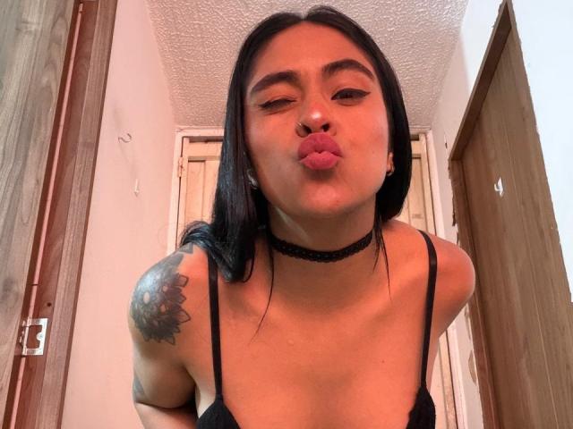 StellaBeckker with black hair, pouty pink lips, black choker, black tank top, tattoo on right shoulder, leaning forward, showing small tits, in a wooden hallway.