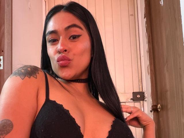 StellaBeckker with long black hair, wearing black lace bra, choker, and pink lipstick, pouting, showing tattoo on right shoulder, holding hair on right side, with cute, sexy look.