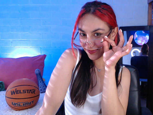AmyStreetwise69 with red and black hair, wearing a white tank top, adjusting her glasses, sitting with a Welsstar basketball on a bed, smiling.