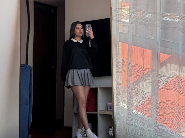 ThaliaAres with long black hair, black sweater, white collar, gray pleated skirt, white socks, holding phone, standing near lace curtain, black TV, and blue shelf in dim room.