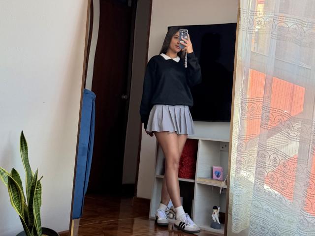 ThaliaAres with long brown hair, black sweater, gray pleated skirt, white socks, and Adidas sneakers, taking a mirror selfie, showing her ass and slender legs.