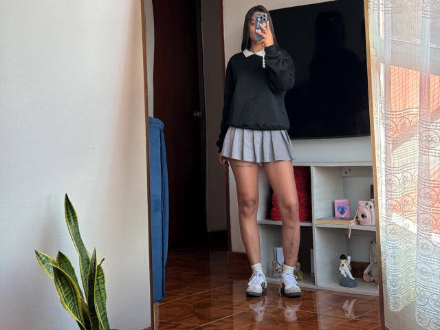 ThaliaAres with medium tits, wearing a black sweater, grey pleated skirt, white socks, and white sneakers, taking a mirror selfie, holding a phone, in a room with a plant.