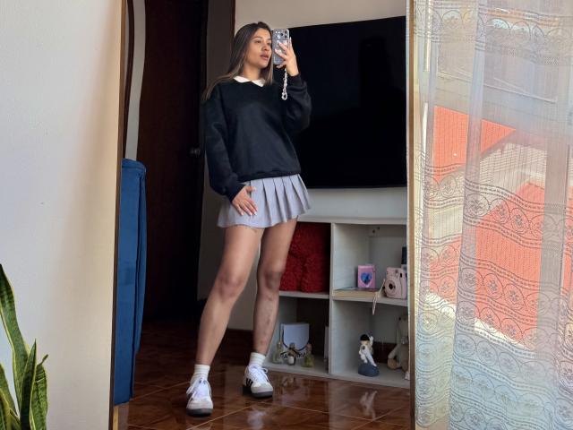 ThaliaAres with long brown hair, black sweater, grey pleated skirt, white socks, and white sneakers, standing in a room, taking a mirror selfie, showing her bare ass.