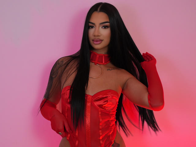 NatalieDelices with long black hair, red satin corset, red gloves, and red choker, holding her hair, showing tattoo on left shoulder, pink and red lighting.