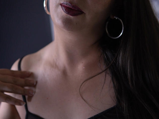 CinthiaBoyer's face and upper chest, showing dark red lipstick, hoop earrings, and a black strap dress. Her hand touches her collarbone, with subtle skin texture visible.