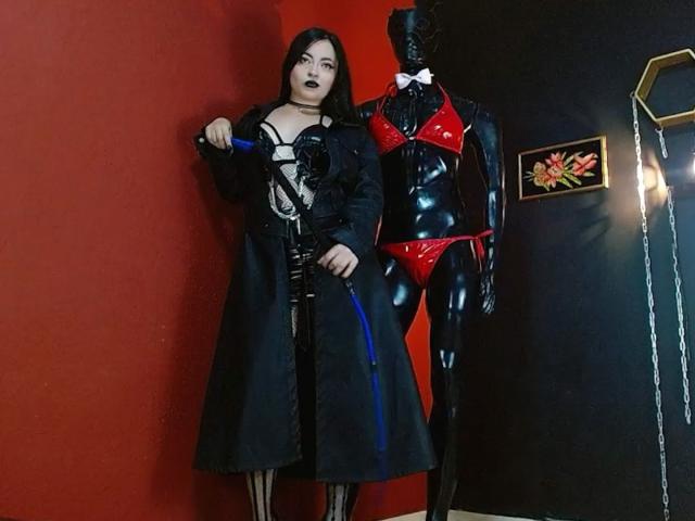 LunaTikka with black hair and dark lipstick, wearing a black lace-up dress and coat, holding a blue leash, next to a shiny black mannequin in red lingerie.