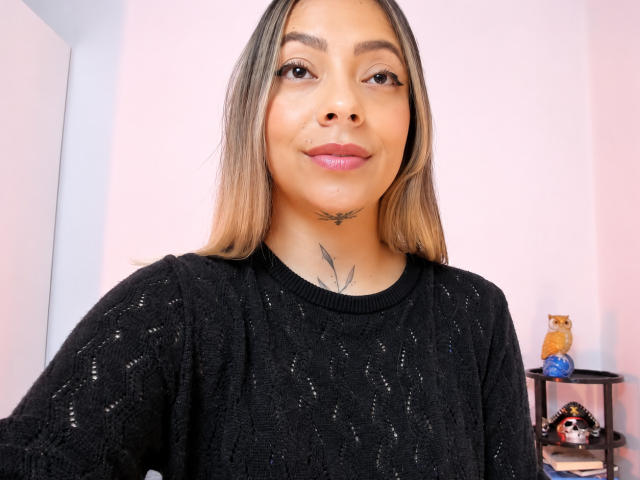 KayleeM with medium skin, brown eyes, and long blonde hair, wearing a black knit sweater, smiling with pink lipstick, small bird tattoo on her neck, and a small shelf in background.