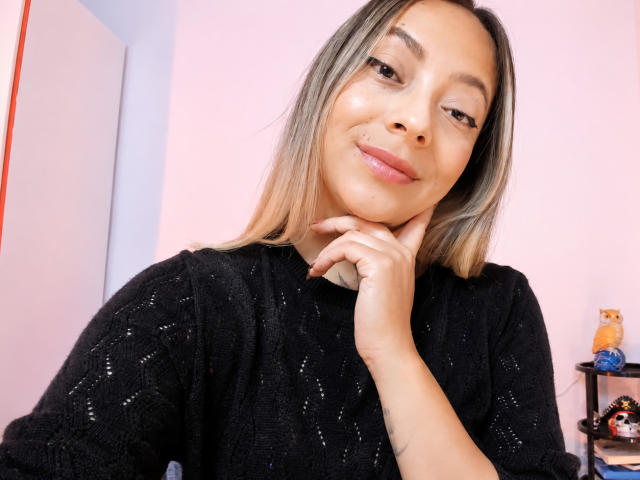 KayleeM with light brown skin, blonde hair, and subtle makeup, wearing a black sweater, smiling slightly, with her hand resting on her chin, showing a cute mole on her cheek.