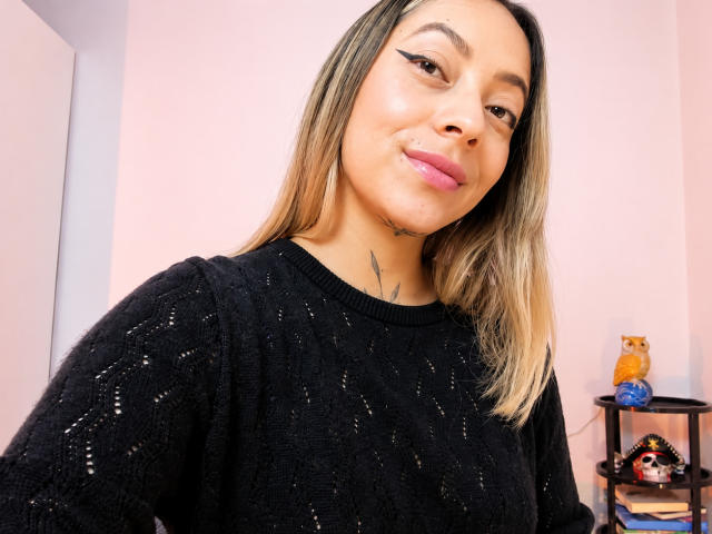 KayleeM with blonde hair, black sparkly sweater, pink lipstick, and small tattoo on neck, smiling at camera, showing her tits slightly.