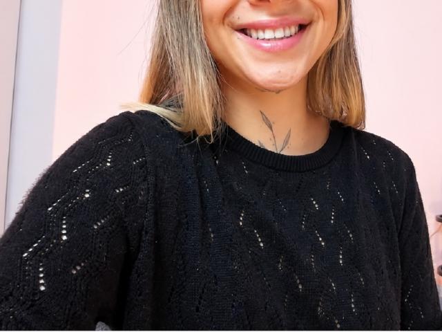 KayleeM, con una sonrisa, lleva un suéter de punto negro, un tatuaje de hoja discreto en el cuello, cabello rubio y un tatuaje en el mentón que se ve ligeramente. Sus senos no son visibles..