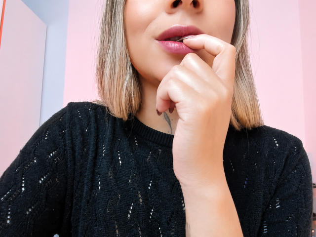 KayleeM's face with her hand near her pink-lipped mouth, wearing a black, sparkly sweater, light brown bob, pink background, and soft, even skin. Boobless, no visible pussy.