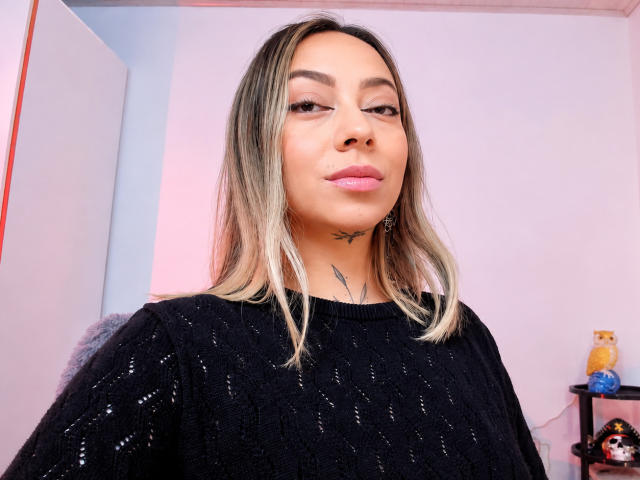 KayleeM with blonde hair, black sweater, pink lips, small black tattoo under chin, slightly raised eyebrow, and subtle makeup, standing in a brightly lit room.