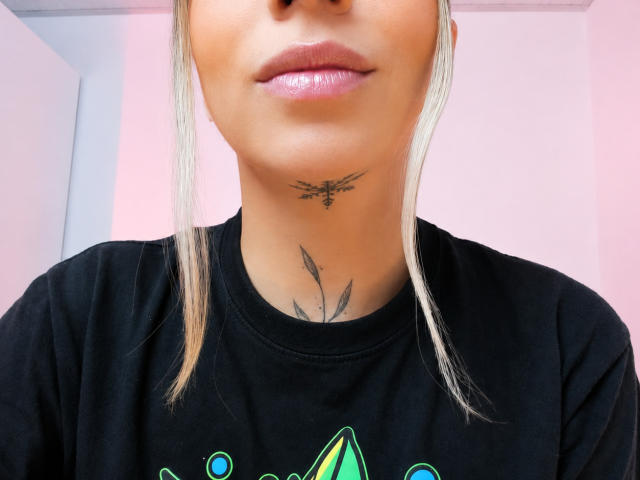 El rostro de KayleeM, con labios rosados brillantes, cabello rubio, camisa negra, un pequeño tatuaje de hoja debajo del mentón y una camisa verde con estampado. Se puede ver un tatuaje en el cuello con un diseño pequeño y delicado..