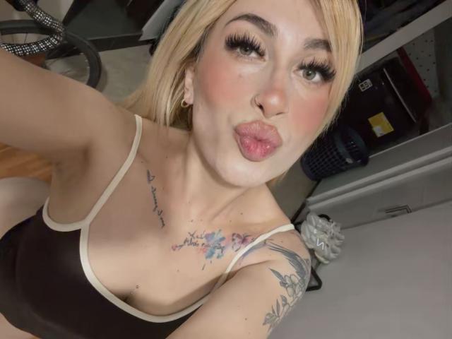SammyViolet with blonde hair, pouty lips, and heavy eyelashes, wearing a black sports bra, showing tattoos on chest and arm, in a selfie with a slight upward angle.