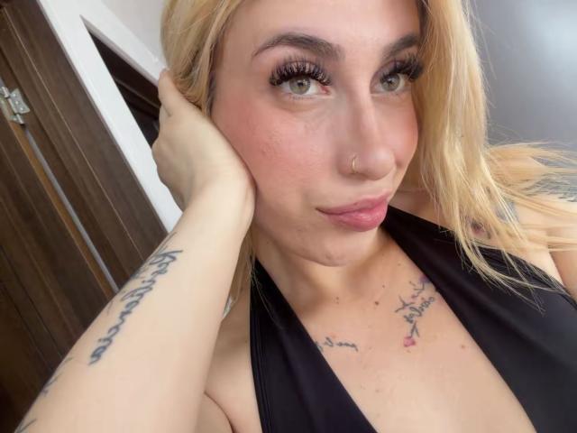 SammyViolet with blonde hair, green eyes, nose ring, and black halter top, showing tattoos on neck and arm, holding face, full lips, and long eyelashes, in selfie.