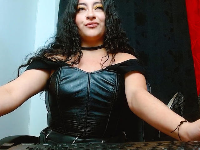 LunaTikka with curly black hair, wearing a black leather off-shoulder top, showing her tits, smiling, arms on a desk, black choker, light skin, confident.