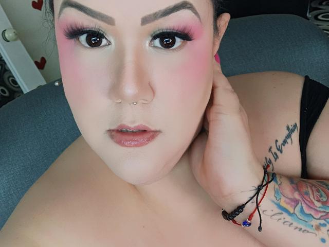 GiiannaMorrys with pink eyeshadow, nose piercing, and tattoo on right wrist, holding neck, wearing black strap, looking at camera with glossy pink lips, blue-gray background.