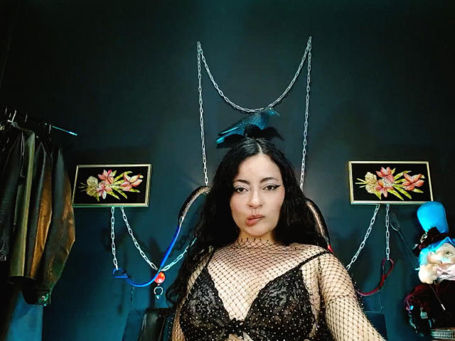 LunaTikka with black fishnet dress, showing her tits, in a room with floral paintings and hanging chains, has long black hair and a nose ring, looking at camera.