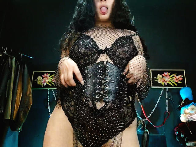 LunaTikka with long black hair, tongue out, wearing a black fishnet top over a lace bodysuit, black corset, and lace-up front, showing her tits and pussy.