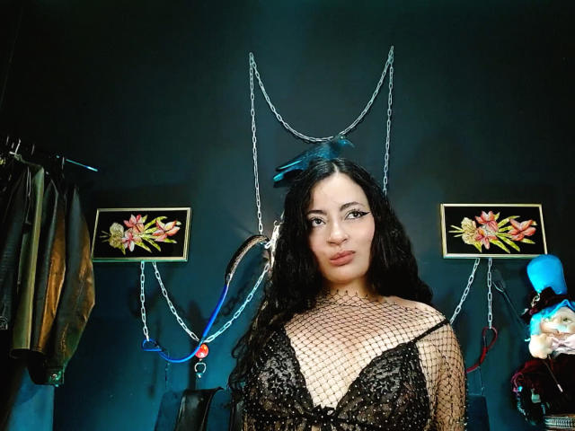 LunaTikka with dark curly hair, wearing a black fishnet top showing her big boobs, standing in front of a dark wall with chains and floral pictures.