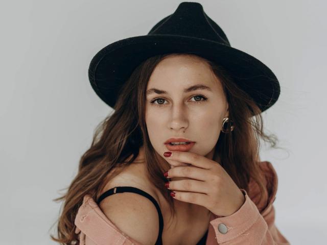 Dilana, con i suoi capelli castani ondulati, indossa un cappello nero, una giacca rosa e orecchini dorati. Ha le unghie rosse, un leggero scollamento e un'espressione sensuale e concentrata..