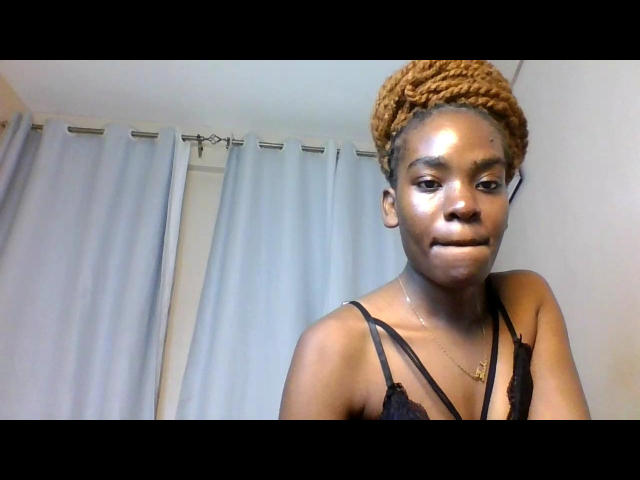 LexyApricot has dark brown skin, small shiny tits, and a braided orange Afro. She wears a black lace top and a gold necklace, smiling slightly.
