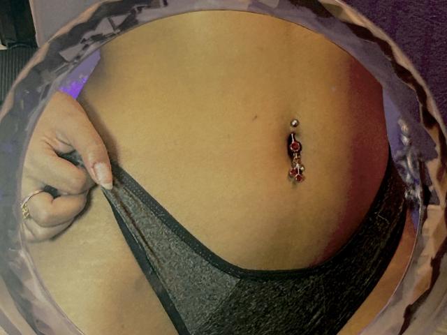 DulceKane's tan belly with a belly button piercing featuring red and gold charms, holding black lace underwear down with her right hand.